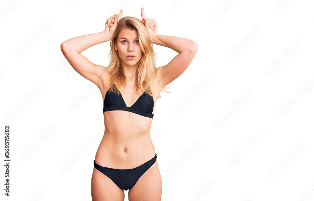 Young beautiful blonde woman wearing bikini doing funny gesture with finger over head as bull horns Young beautiful blonde woman wearing bikini doing funny gesture with finger over head as bull horns