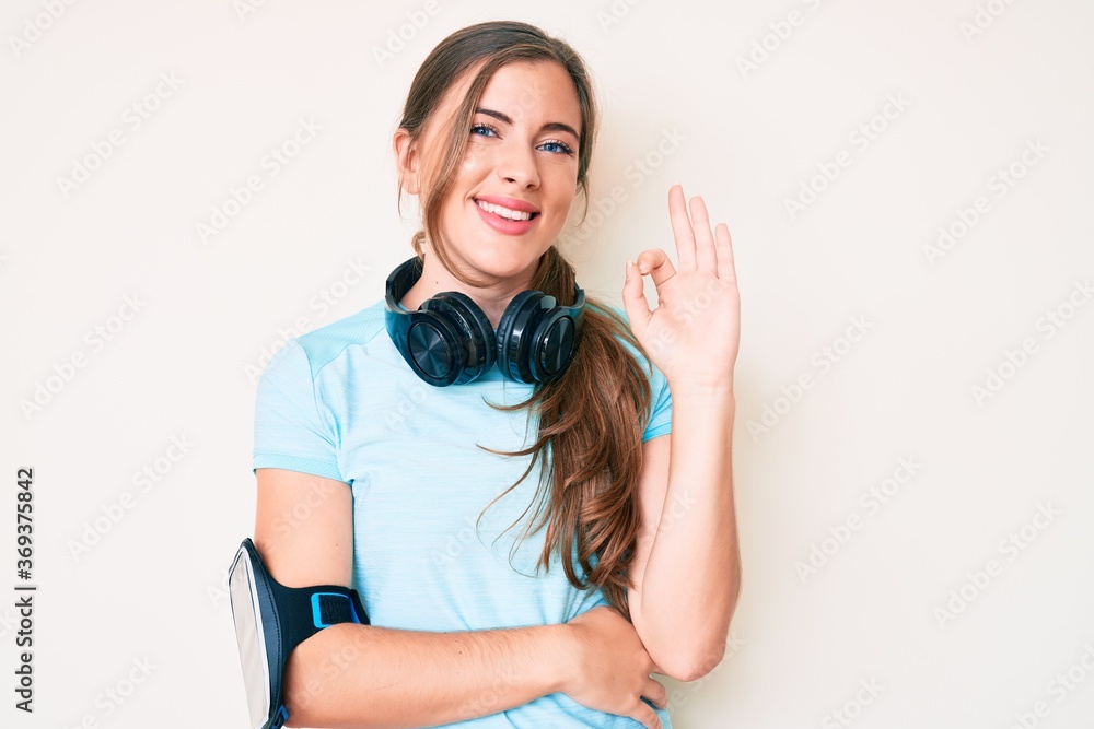 Beautiful young caucasian woman wearing gym clothes and using headphones smiling positive doing ok sign with hand and fingers. successful expression. Beautiful young caucasian woman wearing gym clothes and using headphones smiling positive doing ok sign with hand and fingers. successful expression.