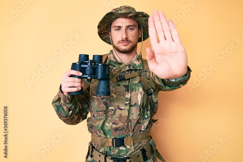 Young caucasian man wearing camouflage army uniform holding binoculars with open hand doing stop sign with serious and confident expression, defense gesture photo