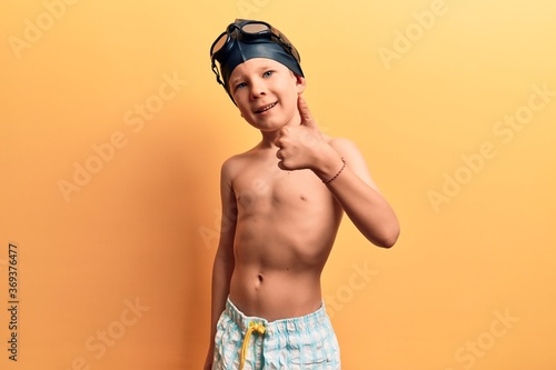 Cute blond kid wearing swimwear and swimmer glasses smiling happy and positive, thumb up doing excellent and approval sign © Krakenimages.com