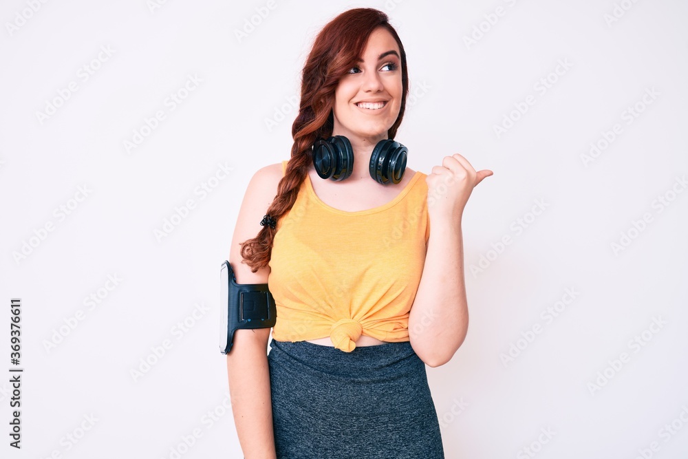 Young beautiful woman wearing gym clothes and using headphones smiling with happy face looking and pointing to the side with thumb up. Young beautiful woman wearing gym clothes and using headphones smiling with happy face looking and pointing to the side with thumb up.