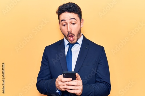 Young hispanic man wearing suit using smartphone scared and amazed with open mouth for surprise, disbelief face © Krakenimages.com