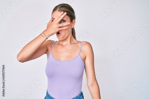 Young blonde girl wearing casual style with sleeveless shirt peeking in shock covering face and eyes with hand, looking through fingers afraid