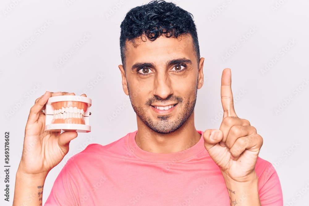 Young hispanic man holding denture smiling with an idea or question pointing finger with happy face, number one Young hispanic man holding denture smiling with an idea or question pointing finger with happy face, number one