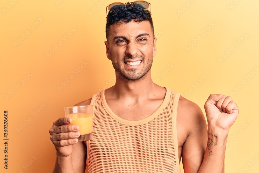 Young hispanic man drinking glass of orange juice screaming proud, celebrating victory and success very excited with raised arm Young hispanic man drinking glass of orange juice screaming proud, celebrating victory and success very excited with raised arm