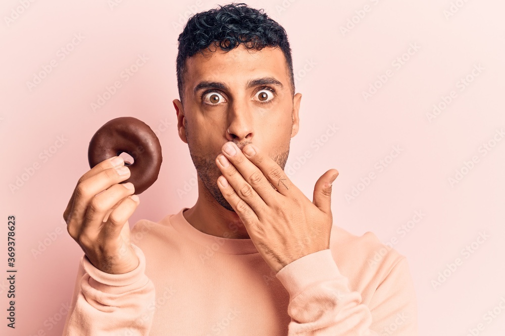 Young hispanic man holding donut covering mouth with hand, shocked and afraid for mistake. surprised expression
