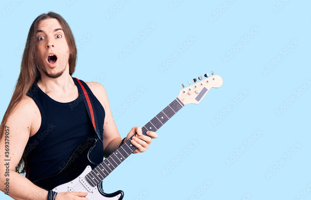 Obraz premium Young adult man with long hair playing electric guitar scared and amazed with open mouth for surprise, disbelief face