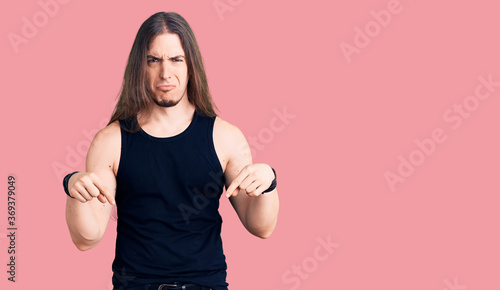 Young adult man with long hair wearing goth style with black clothes pointing down looking sad and upset, indicating direction with fingers, unhappy and depressed.