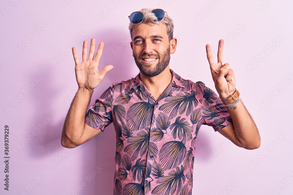 Fototapeta premium Handsome blond man on vacation wearing casual shirt and sunglasses over pink background showing and pointing up with fingers number seven while smiling confident and happy.