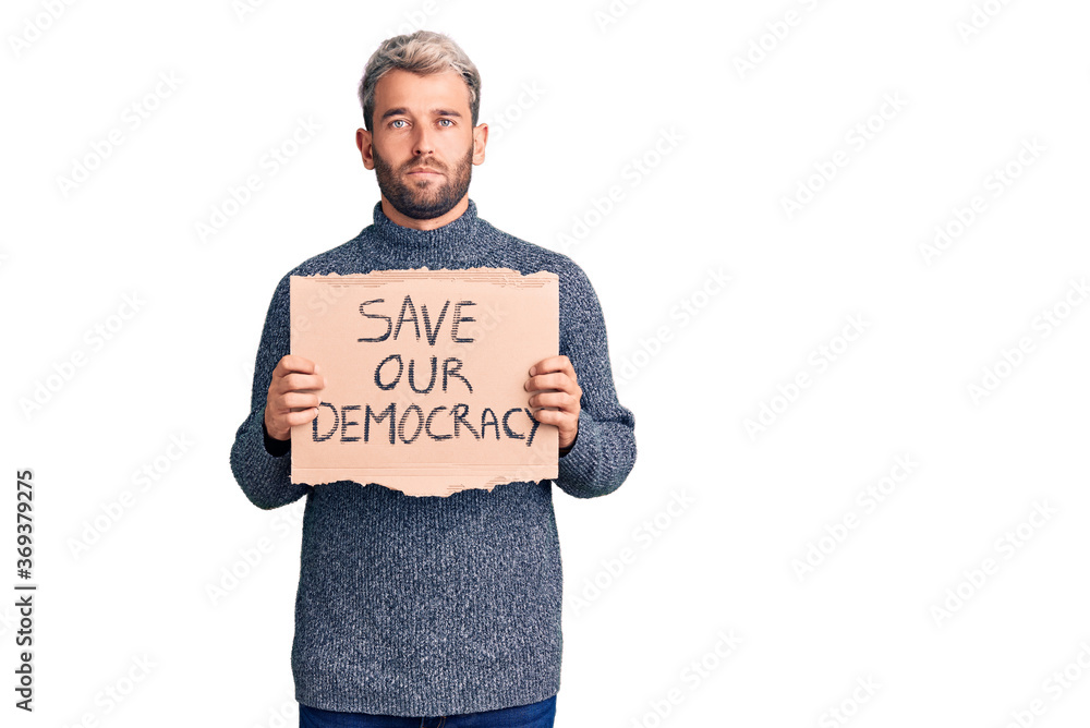 Young blond man holding save our democracy cardboard banner thinking attitude and sober expression looking self confident Young blond man holding save our democracy cardboard banner thinking attitude and sober expression looking self confident