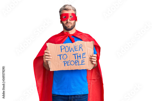 Young blond man wearing super hero custome holding power to the people cardboard banner thinking attitude and sober expression looking self confident