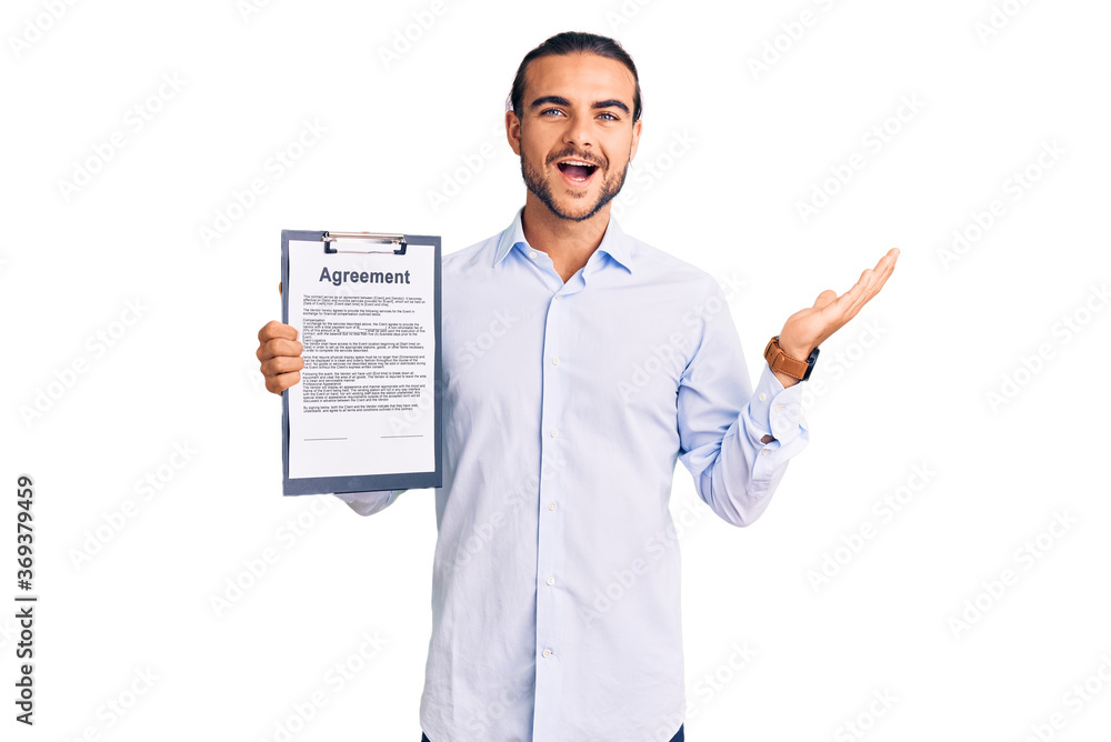 Young handsome man holding clipboard with agreement document celebrating victory with happy smile and winner expression with raised hands Young handsome man holding clipboard with agreement document celebrating victory with happy smile and winner expression with raised hands