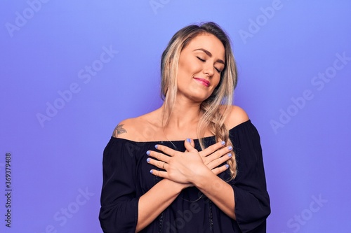 Young beautiful blonde woman wearing casual t-shirt standing over isolated purple background smiling with hands on chest, eyes closed with grateful gesture on face. Health concept.