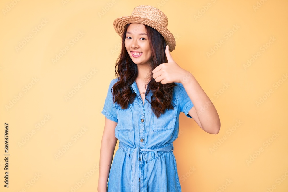Young beautiful chinese girl wearing summer hat doing happy thumbs up gesture with hand. approving expression looking at the camera showing success. Young beautiful chinese girl wearing summer hat doing happy thumbs up gesture with hand. approving expression looking at the camera showing success.