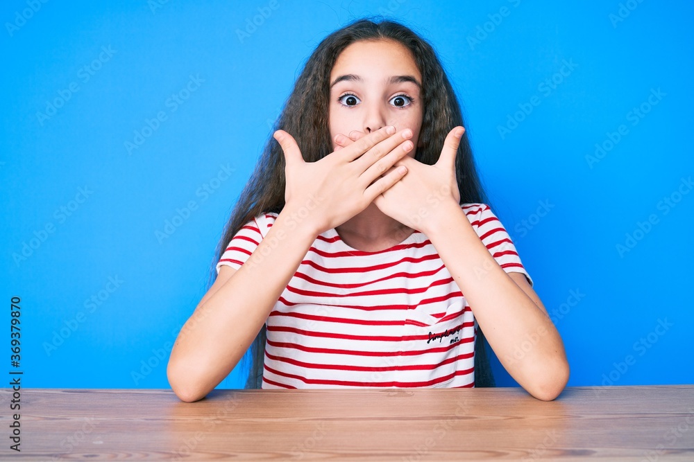 Cute hispanic child girl wearing casual clothes sitting on the table shocked covering mouth with hands for mistake. secret concept. Cute hispanic child girl wearing casual clothes sitting on the table shocked covering mouth with hands for mistake. secret concept.