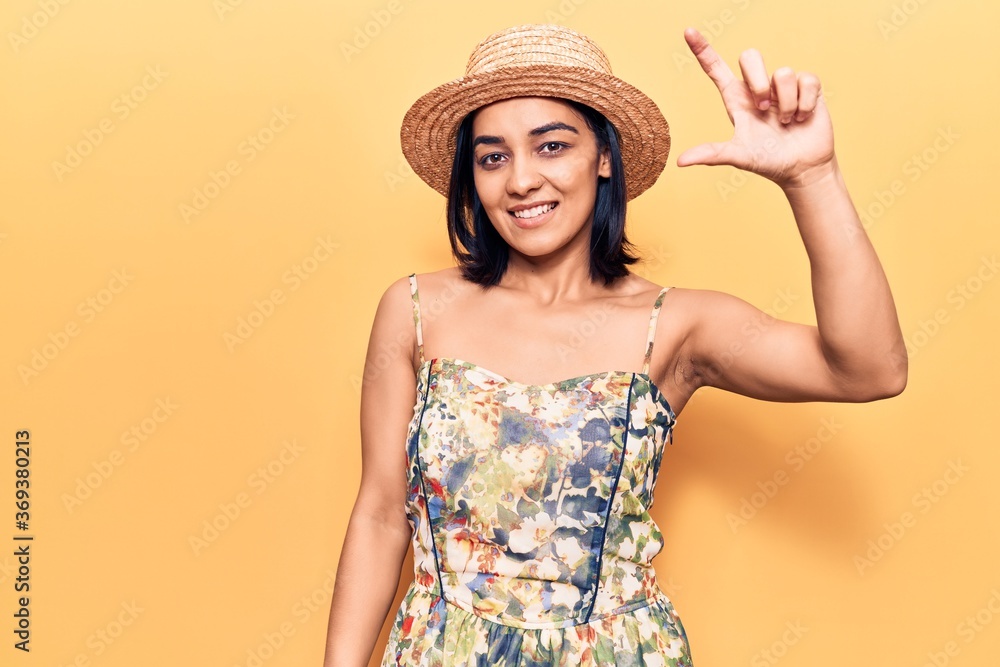 Young beautiful latin woman wearing summer hat smiling and confident gesturing with hand doing small size sign with fingers looking and the camera. measure concept. Young beautiful latin woman wearing summer hat smiling and confident gesturing with hand doing small size sign with fingers looking and the camera. measure concept.