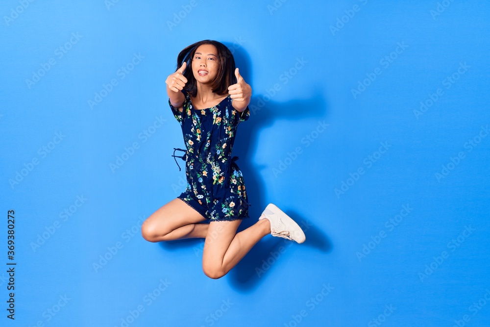 Young beautiful chinese woman wearing casual clothes smiling happy. Jumping with smile on face doing ok sign with thumbs up over isolated blue background