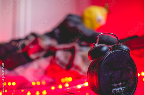 Classic black alarm clock from behind with red lights and boy with lemon mask out of focus behind