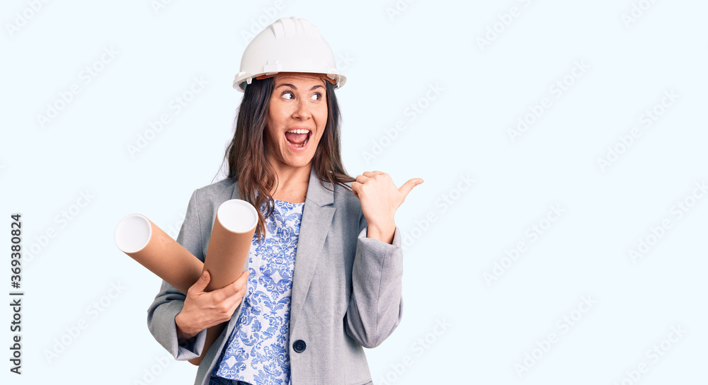 Young beautiful brunette woman wearing architect hardhat holding blueprint pointing thumb up to the side smiling happy with open mouth Young beautiful brunette woman wearing architect hardhat holding blueprint pointing thumb up to the side smiling happy with open mouth