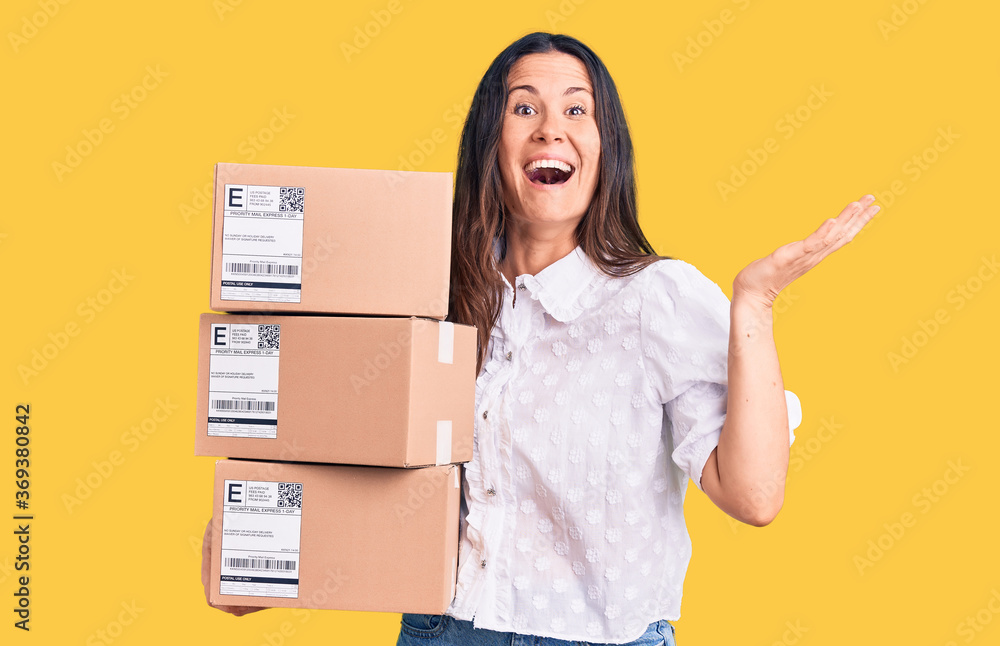 Young beautiful brunette woman holding delivery cardboard package celebrating victory with happy smile and winner expression with raised hands Young beautiful brunette woman holding delivery cardboard package celebrating victory with happy smile and winner expression with raised hands