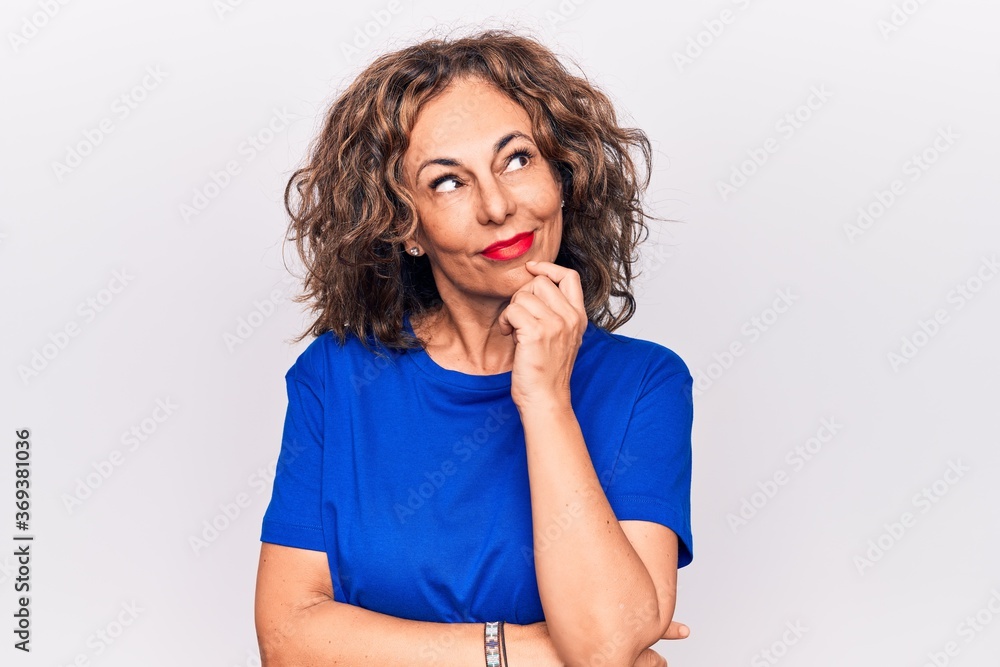 Middle age beautiful brunette woman wearing blue t-shirt standing over white background thinking concentrated about doubt with finger on chin and looking up wondering Middle age beautiful brunette woman wearing blue t-shirt standing over white background thinking concentrated about doubt with finger on chin and looking up wondering