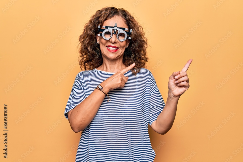 Middle age beautiful woman controlling vision using optometry glasses over yellow background smiling and looking at the camera pointing with two hands and fingers to the side. Middle age beautiful woman controlling vision using optometry glasses over yellow background smiling and looking at the camera pointing with two hands and fingers to the side.