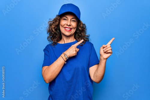 Middle age beautiful delivery woman wearing blue uniform and cap over isolated background smiling and looking at the camera pointing with two hands and fingers to the side.