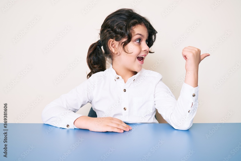 Cute hispanic child wearing casual clothes sitting on the table pointing thumb up to the side smiling happy with open mouth