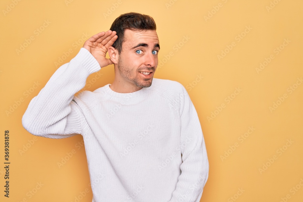 Young handsome man with blue eyes wearing casual sweater standing over yellow background smiling with hand over ear listening an hearing to rumor or gossip. Deafness concept. Young handsome man with blue eyes wearing casual sweater standing over yellow background smiling with hand over ear listening an hearing to rumor or gossip. Deafness concept.