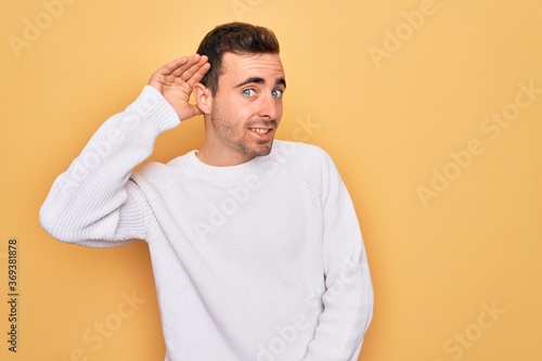 Young handsome man with blue eyes wearing casual sweater standing over yellow background smiling with hand over ear listening an hearing to rumor or gossip. Deafness concept. © Krakenimages.com