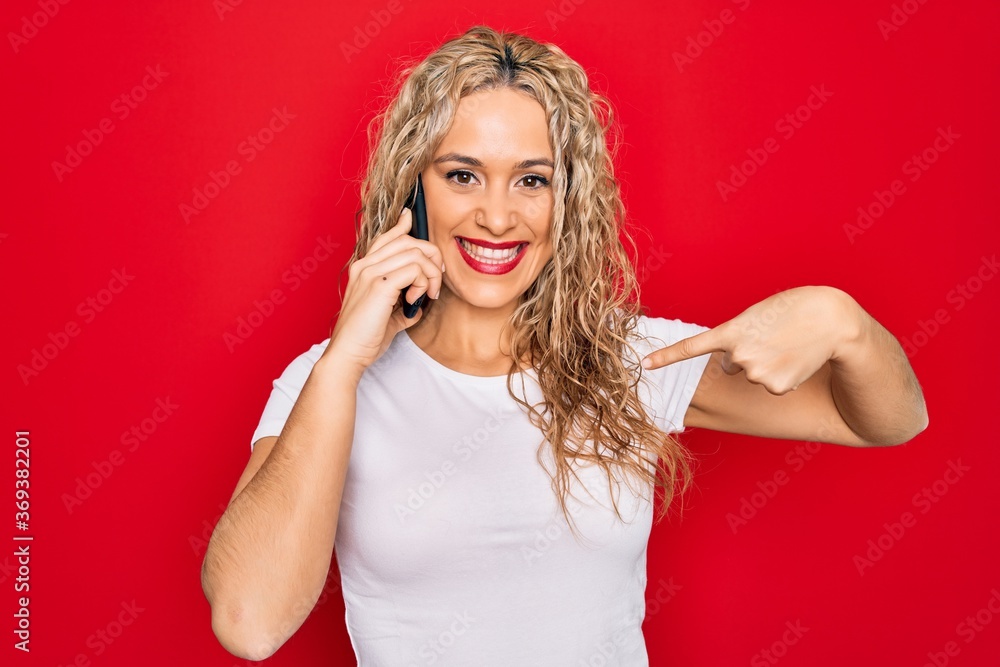 Beautiful blonde woman having conversation talking on the smartphone over red background pointing finger to one self smiling happy and proud