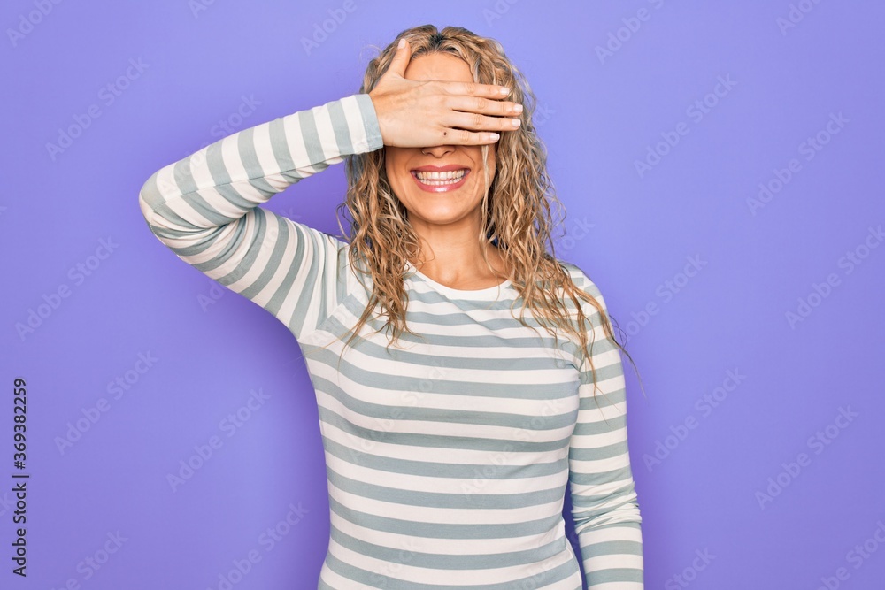 Beautiful blonde woman wearing casual striped t-shirt and glasses over purple background smiling and laughing with hand on face covering eyes for surprise. Blind concept. Beautiful blonde woman wearing casual striped t-shirt and glasses over purple background smiling and laughing with hand on face covering eyes for surprise. Blind concept.