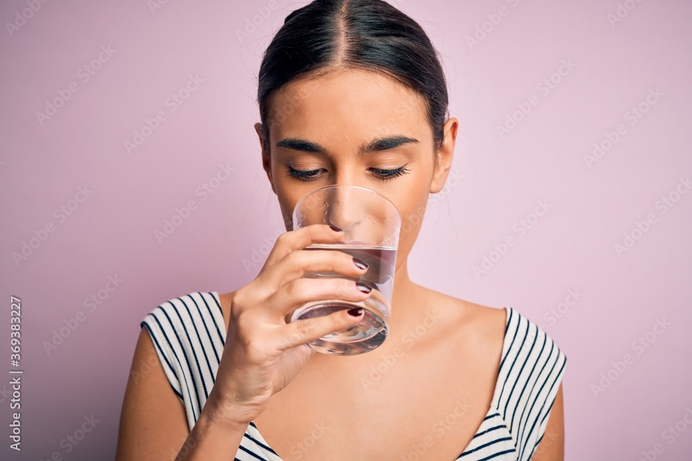 Young beautiful brunette woman drinking glass of healthy water to refreshment standing over isolated pink background Young beautiful brunette woman drinking glass of healthy water to refreshment standing over isolated pink background