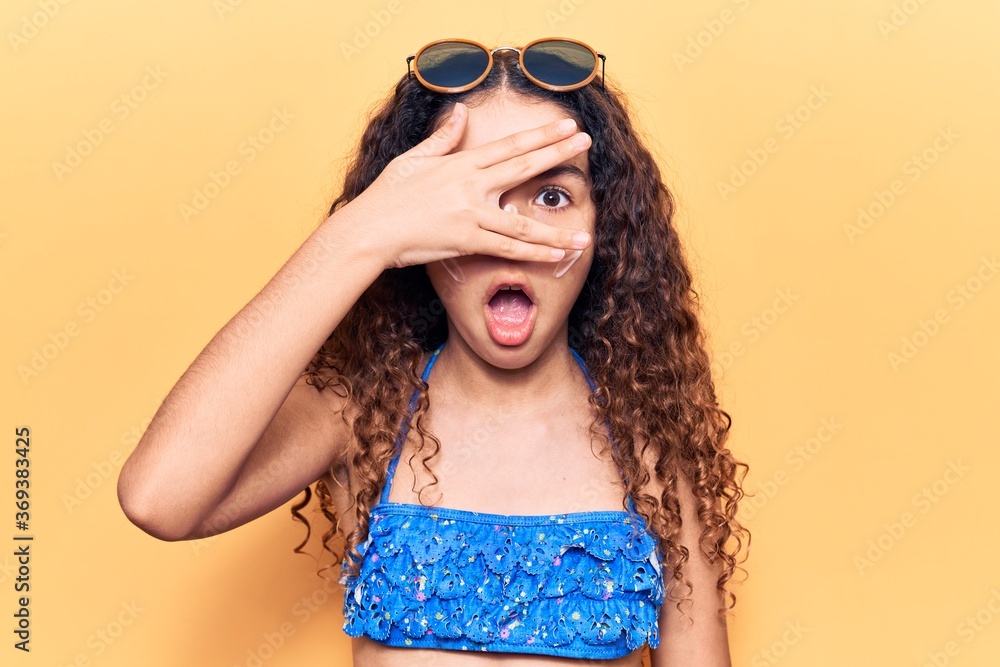 Beautiful kid girl with curly hair wearing bikini and sunglasses peeking in shock covering face and eyes with hand, looking through fingers afraid Beautiful kid girl with curly hair wearing bikini and sunglasses peeking in shock covering face and eyes with hand, looking through fingers afraid