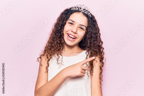 Beautiful kid girl with curly hair wearing princess tiara smiling cheerful pointing with hand and finger up to the side
