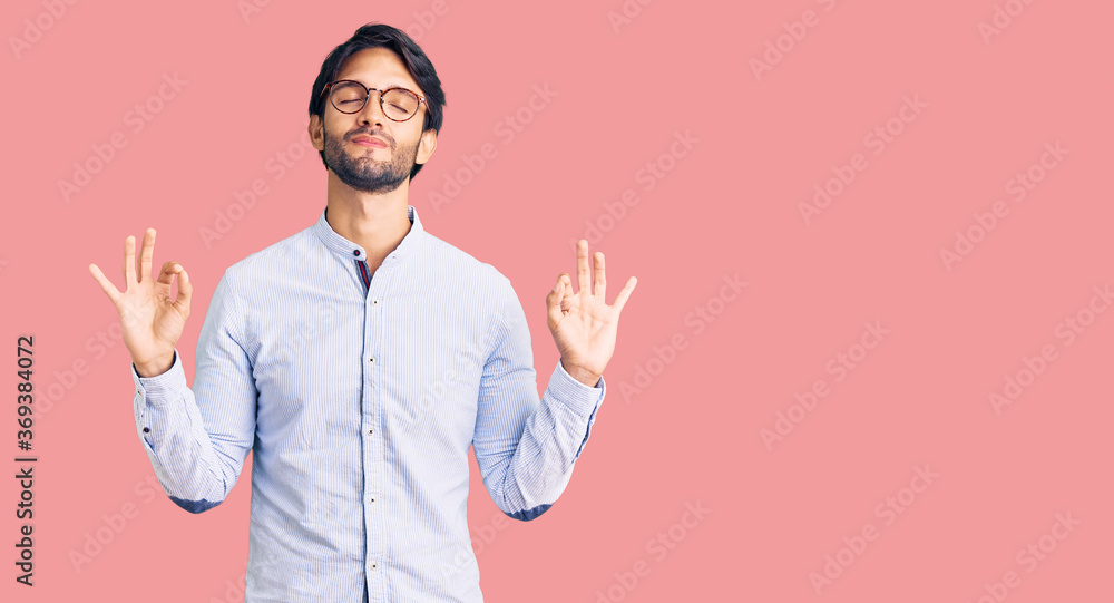 Handsome hispanic man wearing business shirt and glasses relaxed and smiling with eyes closed doing meditation gesture with fingers. yoga concept.
