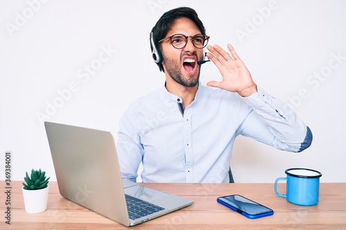Handsome hispanic man working at the office wearing operator headset shouting and screaming loud to side with hand on mouth. communication concept.