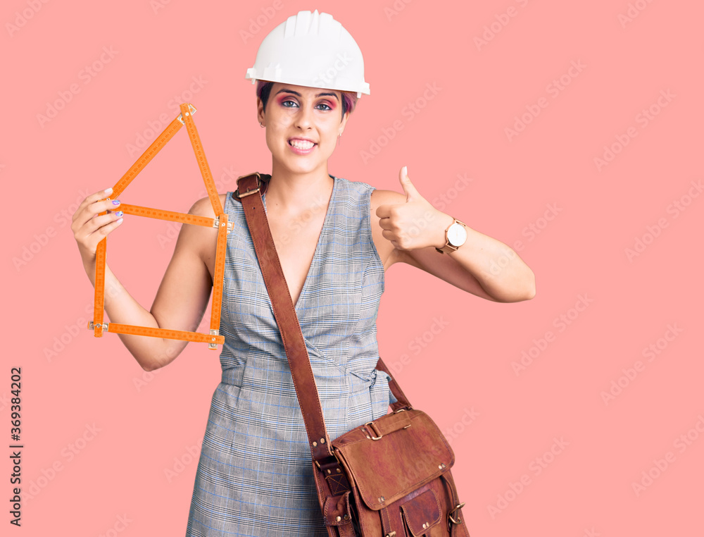 Young beautiful woman wearing architect hardhat holding build project smiling happy and positive, thumb up doing excellent and approval sign Young beautiful woman wearing architect hardhat holding build project smiling happy and positive, thumb up doing excellent and approval sign