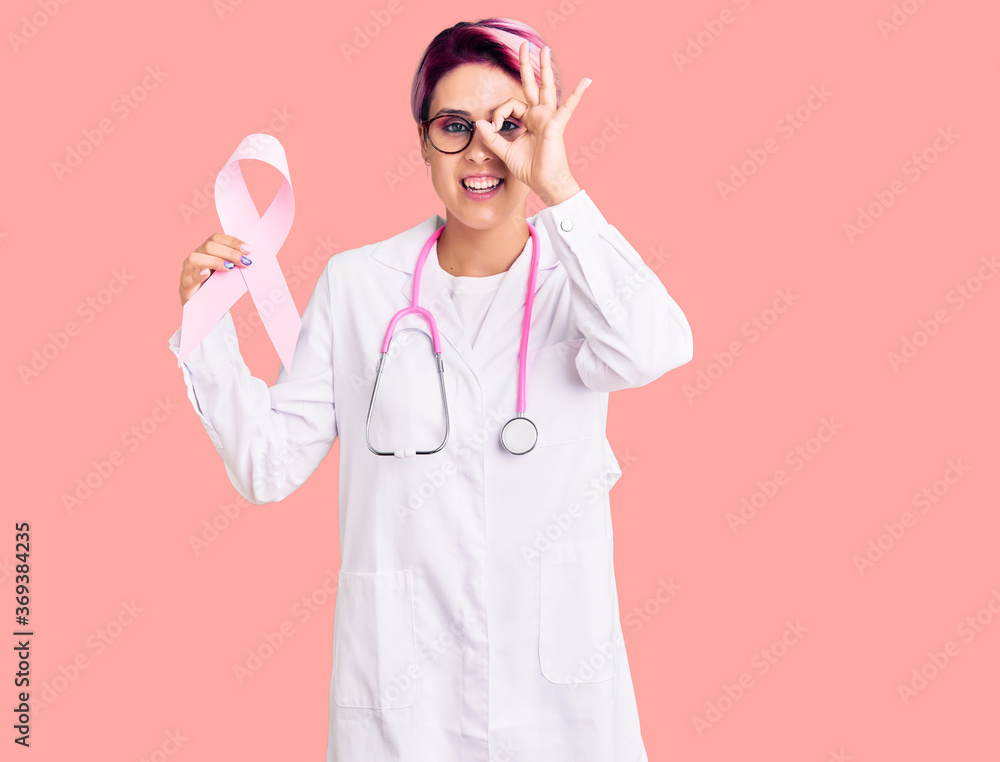 Young beautiful woman with pink hair wearing doctor stethoscope holding cancer ribbon smiling happy doing ok sign with hand on eye looking through fingers Young beautiful woman with pink hair wearing doctor stethoscope holding cancer ribbon smiling happy doing ok sign with hand on eye looking through fingers