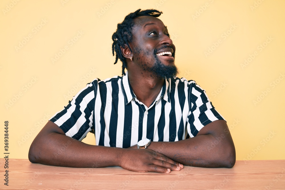 Young african american man with braids wearing casual clothes sitting on the table looking away to side with smile on face, natural expression. laughing confident. Young african american man with braids wearing casual clothes sitting on the table looking away to side with smile on face, natural expression. laughing confident.