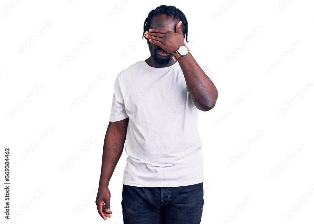 Young african american man with braids wearing casual white tshirt covering eyes with hand, looking serious and sad. sightless, hiding and rejection concept Young african american man with braids wearing casual white tshirt covering eyes with hand, looking serious and sad. sightless, hiding and rejection concept