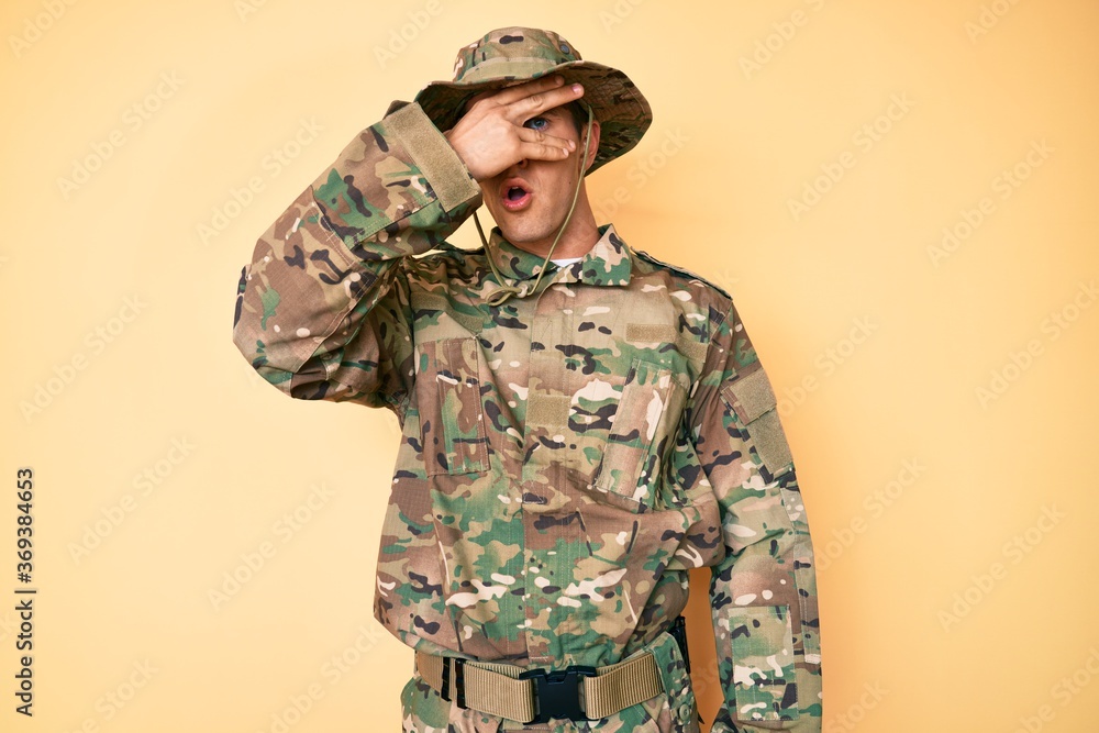 Young handsome man wearing camouflage army uniform peeking in shock covering face and eyes with hand, looking through fingers with embarrassed expression. Young handsome man wearing camouflage army uniform peeking in shock covering face and eyes with hand, looking through fingers with embarrassed expression.