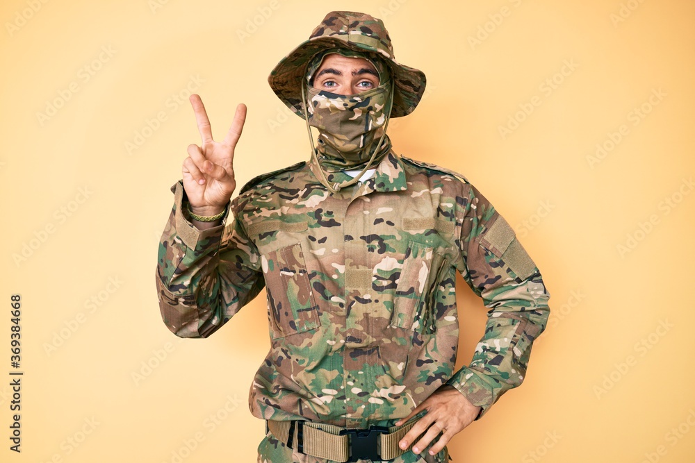 Young handsome man wearing camouflage army uniform and balaclava smiling with happy face winking at the camera doing victory sign. number two. Young handsome man wearing camouflage army uniform and balaclava smiling with happy face winking at the camera doing victory sign. number two.