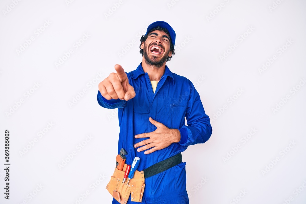 Handsome young man with curly hair and bear weaing handyman uniform laughing at you, pointing finger to the camera with hand over body, shame expression Handsome young man with curly hair and bear weaing handyman uniform laughing at you, pointing finger to the camera with hand over body, shame expression