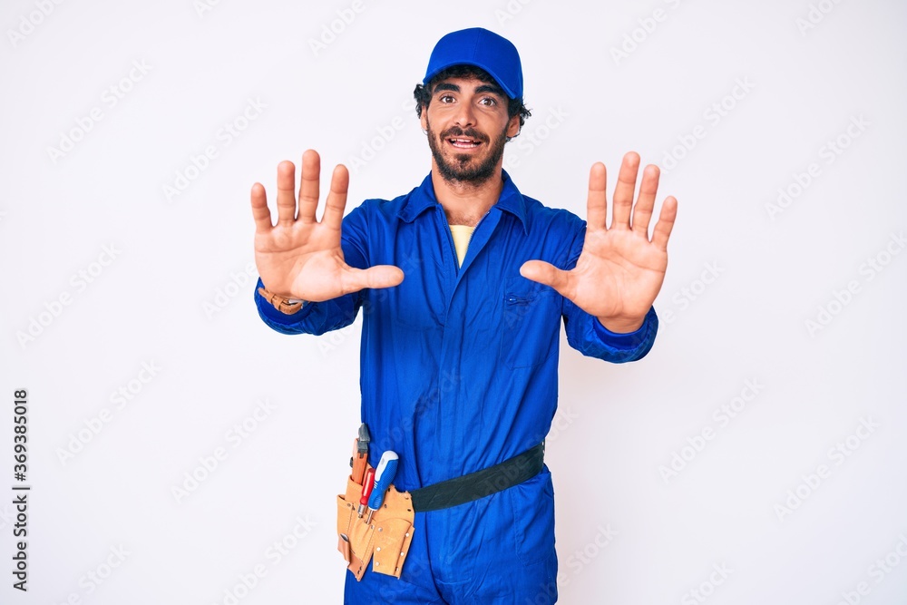 Handsome young man with curly hair and bear weaing handyman uniform doing stop gesture with hands palms, angry and frustration expression