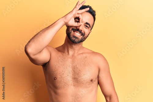 Young hispanic man standing shirtless smiling happy doing ok sign with hand on eye looking through fingers