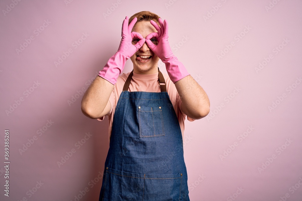 Young handsome redhead cleaner man doing housework wearing apron and gloves doing ok gesture like binoculars sticking tongue out, eyes looking through fingers. Crazy expression. Young handsome redhead cleaner man doing housework wearing apron and gloves doing ok gesture like binoculars sticking tongue out, eyes looking through fingers. Crazy expression.