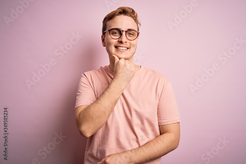 Young handsome redhead man wearing casual t-shirt standing over isolated pink background looking confident at the camera smiling with crossed arms and hand raised on chin. Thinking positive.