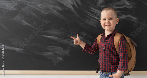 A young schoolboy near the blackboard shows his index finger to the left. A fashionable guy is wearing a plaid shirt and light jeans. On the back of the child is a brown backpack. Education in