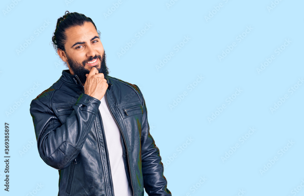 Young arab man wearing casual leather jacket looking confident at the camera smiling with crossed arms and hand raised on chin. thinking positive. Young arab man wearing casual leather jacket looking confident at the camera smiling with crossed arms and hand raised on chin. thinking positive.